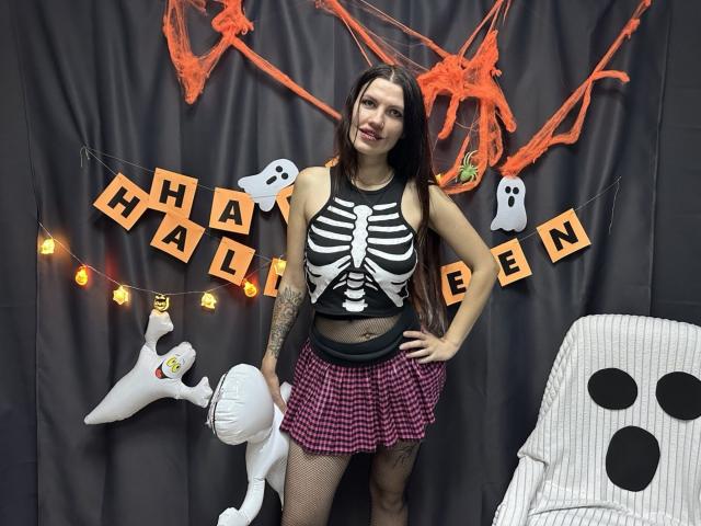MilaLoona with long brown hair, wearing a black skeleton top, red checkered skirt, and fishnet stockings, standing next to a white ghost, showing her tattooed arm and smiling.