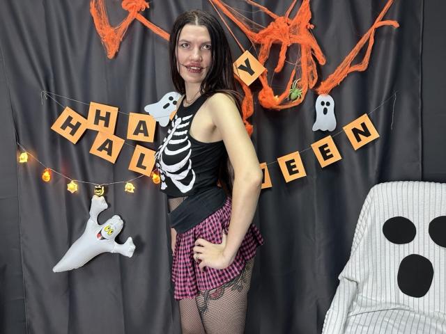 MilaLoona with long brown hair, black skeleton top, pink checkered skirt, fishnet stockings, smiling, standing, Halloween decor, orange and black, ghost, spooky, grey curtain, chair.