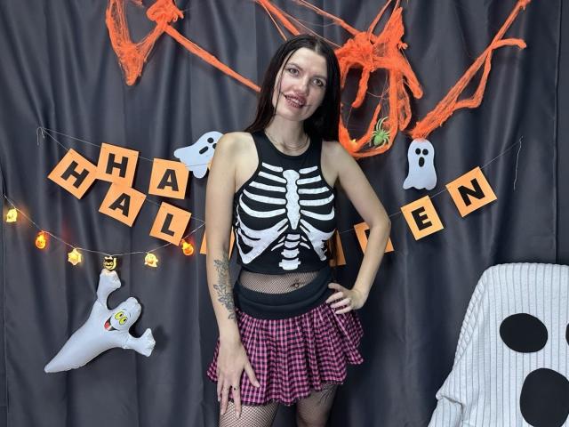 MilaLoona with long brown hair, smiling, wearing a skeleton crop top, red checkered skirt, fishnet stockings, and tattoos, standing in front of Halloween decorations. Boobs visible under top.