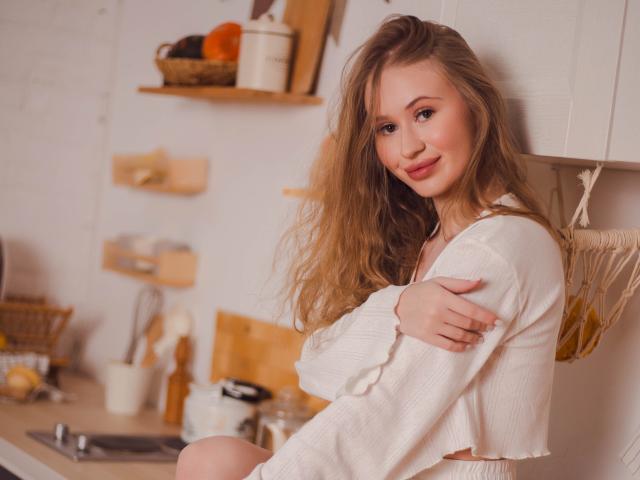 ViolettaSteward with long blonde hair, smiling, wearing a white cropped shirt, arms crossed, showing her tits, in a cozy kitchen. Soft light, relaxed, cute.