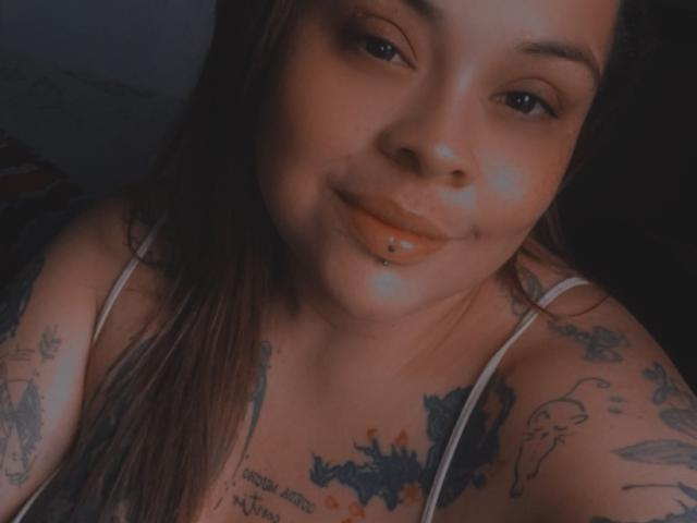Diianii with dark brown hair, smiling with a labret piercing, tattoos on her shoulders and chest, wearing a white strap top, in a dimly lit selfie.
