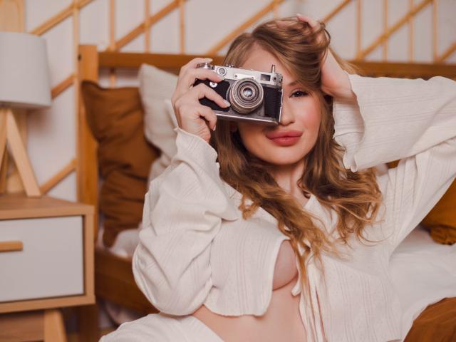 ViolettaSteward with wavy blonde hair, holding a vintage camera, wearing an open white shirt, exposing her tits, lying in a wooden bed with brown pillows, smiling.