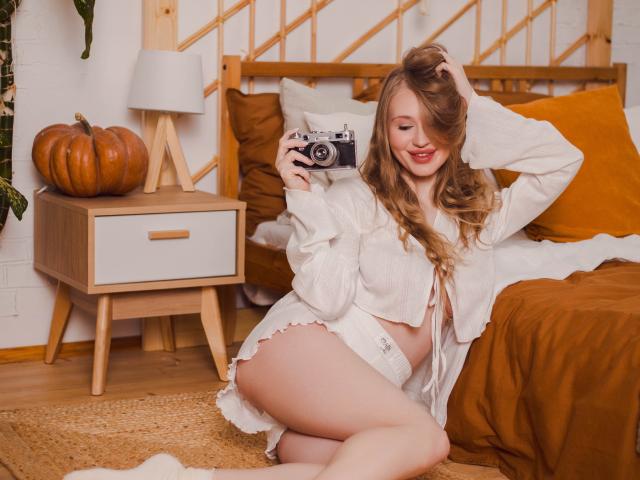 ViolettaSteward with wavy blonde hair, red lipstick, white shirt, and white panties, kneeling, holding a camera, smiling, showing her ass, in a cozy bedroom.