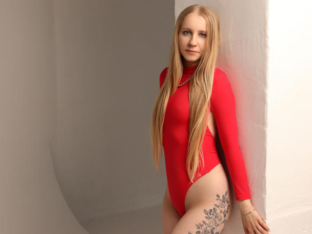 MilanaSun with long blonde hair wearing a red, long-sleeve, high-cut one-piece, showing her ass and a large floral tattoo on her right thigh, leaning against a white wall.
