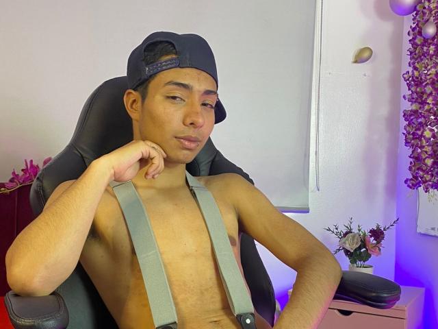DiiGiacomo, senza camicia, indossa bretelle grigie e un berretto nero da baseball, con un'espressione compiaciuta, con il mento appoggiato sulla mano, seduto su una sedia da gaming nera..