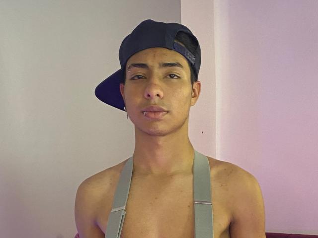 DiiGiacomo is a young dude with tan skin, wearing a backwards navy cap and gray suspenders, looking at the camera with his tongue slightly out, shirtless.