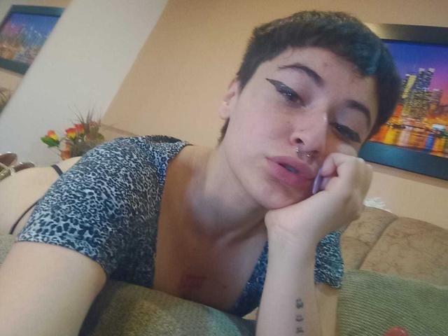 MissTat with short dark hair, black eyeliner, nose ring, and lip piercing, wearing a leopard print top, resting her chin on her hand, showing her tits and tattoo.