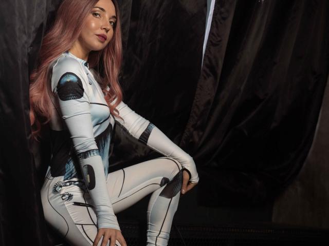 LettieSweet, with pink hair, wearing a tight white and black sci-fi bodysuit, sits against a dark curtain, showing her small ass and white boots, looking serious.