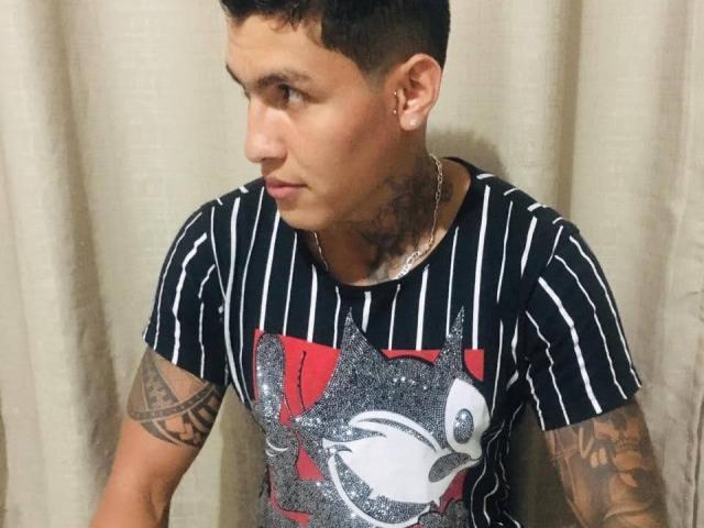 AbrahamStiff with short curly hair, tattoos on neck and arm, wearing a black and white striped shirt with a graphic, looking to the side, silver chain necklace.