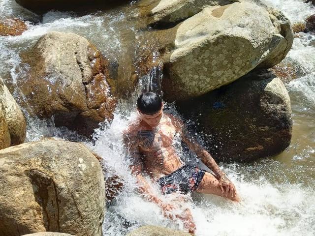 AbrahamStiff, shirtless with black tattoos, sits in rocky stream, water splashing over his chest and legs, wearing black and red swim trunks, wet skin, dark hair.