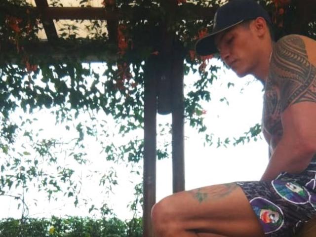 AbrahamStiff, shirtless with tattoos, wearing a blue cap and patterned shorts, sitting under a wooden pergola with leafy vines, showing a tattoo on his thigh.