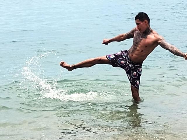 AbrahamStiff, shirtless with tattoos, kicking up water in a blue ocean, wearing purple and white patterned shorts, right leg extended high, splashing.