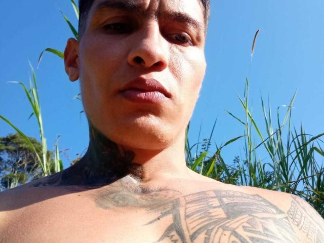AbrahamStiff's shirtless face with a tattoo on his neck and chest, short dark hair, and a neutral expression under a bright blue sky with tall green grass.
