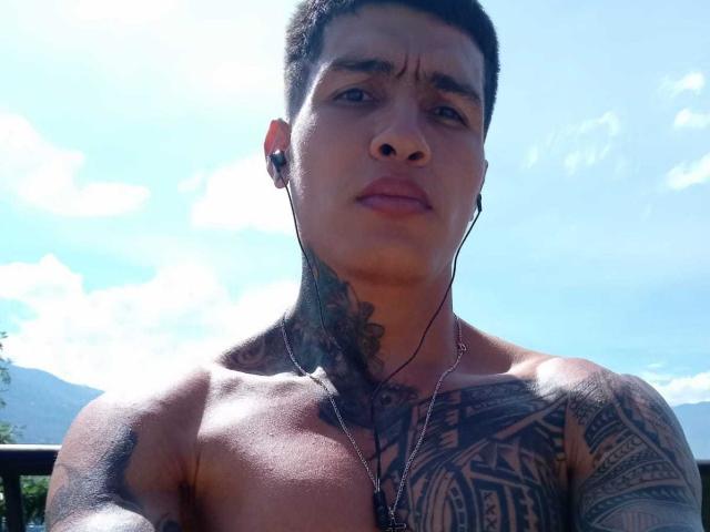 AbrahamStiff with short black hair, tattoos on neck and chest, earbuds in, shirtless, looking slightly confused, sunlight on skin, blue sky in background.
