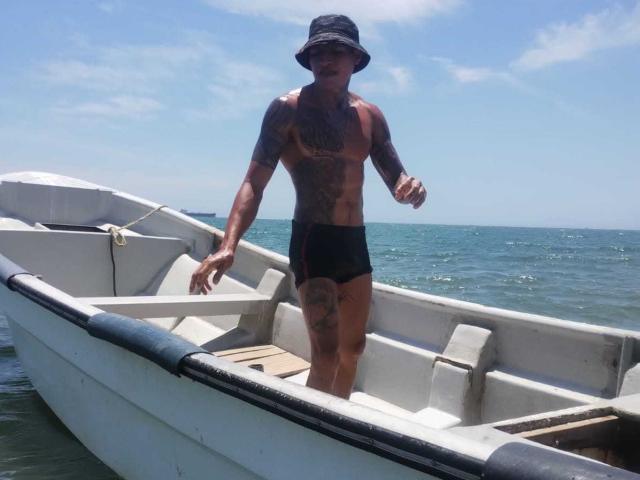 Shirtless AbrahamStiff with tattoos, wearing black shorts and a bucket hat, stands in a white boat on blue water, looking forward with a slight smile.