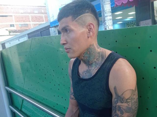 AbrahamStiff with a shaved side and back, black tank top, tattoos on neck and arm, looking to the side, standing against a green perforated wall.
