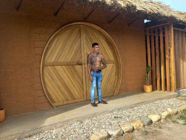 AbrahamStiff stands shirtless with tattoos, wearing blue jeans and black shoes, in front of a wooden circular door, against a brown adobe wall with a thatched roof.