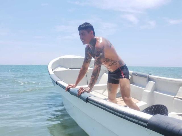 AbrahamStiff, tattooed and shirtless, leans on a white boat's edge, wearing black speedo with red stripes, water's blue-green, clear sky, sunlight, black hair, muscular, standing, hands gripping.