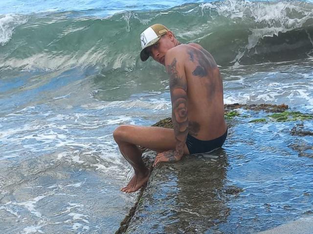 AbrahamStiff, shirtless with tattoos, wearing a baseball cap and black shorts, sits on a wet rock in the ocean with waves crashing around him.