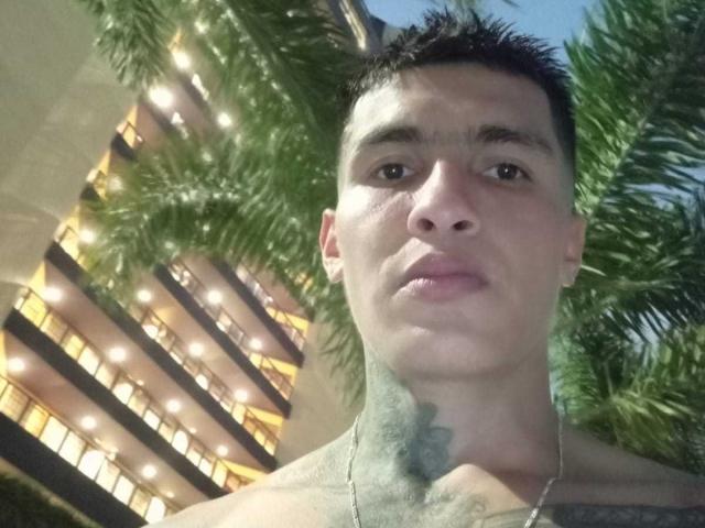 AbrahamStiff with short black hair, tattoo on neck, shirtless, looking up, standing in front of palm tree, building with lit windows in background, clear blue sky.