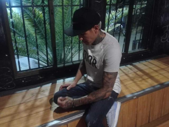 AbrahamStiff sits on a wooden ledge, wearing a black cap, white t-shirt, and torn jeans, with tattooed arms and a black bracelet, looking down at his hands.