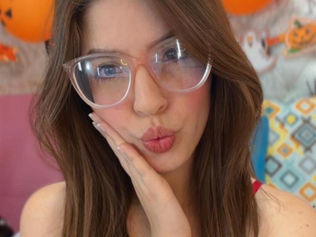 NatyBakers met bruin haar, heldere brillen en roze lippen, die een frons trekt, met een hand op de wang, gekleed in een rode top, reflectie in de brillen, zachte roze blush, lichte huid, schattig gezicht..