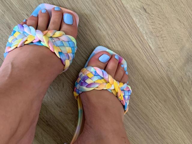 Babycamy-hot's feet with colorful, woven sandals on a wooden floor. She's got blue toenails, and her toes are slightly curled. Her sandals are rainbow-patterned with yellow, purple, and blue.