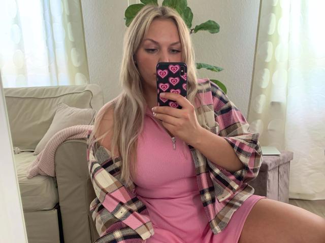 Babycamy, con i capelli biondi, un vestito rosa e una camicia a quadri, sta scattando un selfie allo specchio, mostrando il seno nel riflesso, e tiene in mano un telefono con motivi a cuori rosa..
