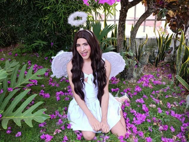 FlorenceLambert with long brown hair, white dress, and angel wings, kneeling on grass with purple flowers, smiling, wearing a halo, in a garden. Boobs are small, tits are cute.