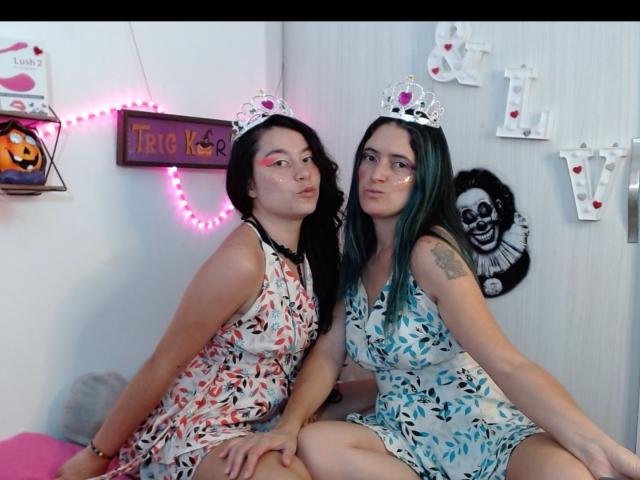 LunaVenus with black hair and pink face paint, wearing a floral dress, sits next to LunaVenus with green hair, wearing a blue floral dress, both with silver tiaras.