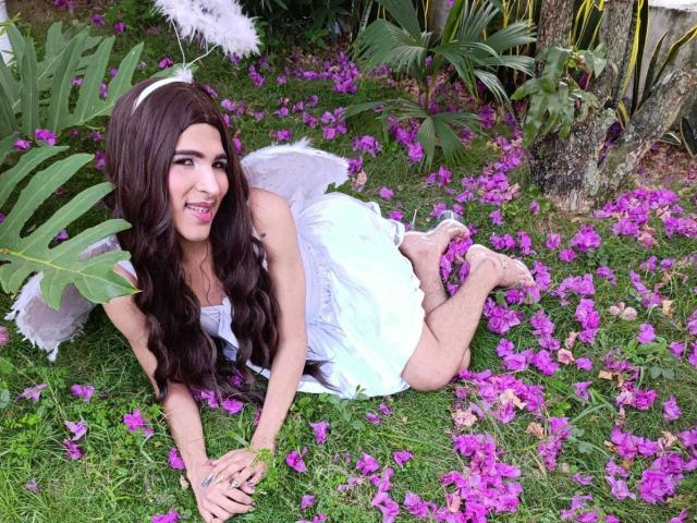 FlorenceLambert, with long brown hair, white dress, and small white wings, lies on grass, surrounded by purple flowers, smiling, showing her ass, and wearing a white headband.