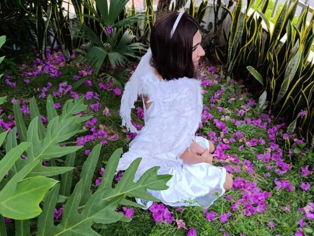 FlorenceLambert, with white angel wings and a white dress, kneels in purple flowers, surrounded by green leaves, has dark brown hair and a white headband.