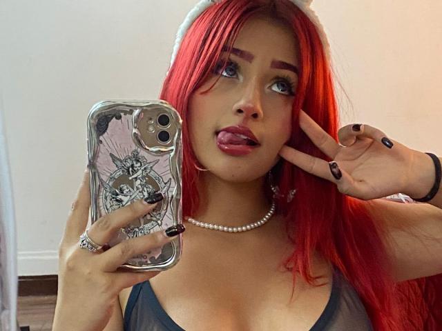 AlanaOdinsson, de pelo pelirrojo, con la lengua afuera, vistiendo un collar de perlas, sosteniendo un teléfono con una funda con forma de calavera, haciendo un gesto de la paz, mostrando sus pechos con una blusa transparente..