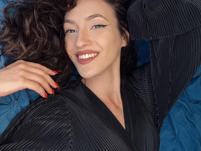 VievienneClemente with curly brown hair, smiling with red lipstick, black pinstripe shirt, and red nails, lying on a blue surface, arm raised, showing her tits.
