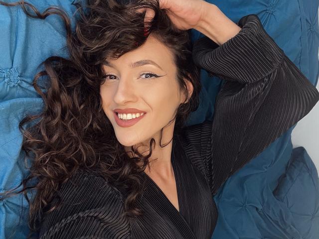 VievienneClemente with curly brown hair, smiling, wearing a black pinstripe shirt, lying on a blue quilted blanket, hand in hair, bold eyeliner, and pink lipstick.