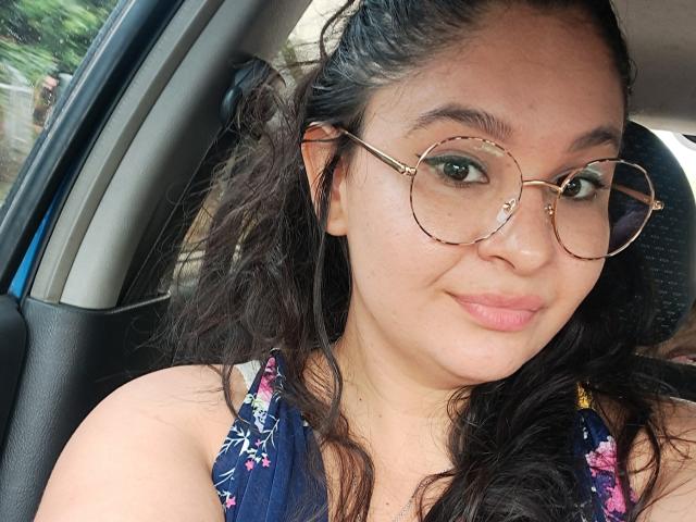 ArianneHot with curly black hair, wearing round gold glasses, and a floral blue top, sitting in a car, smiling with pink lipstick, showing her cute, curvy face.