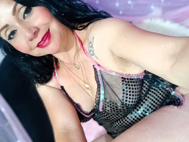 Zaria69 with black hair, red lipstick, wearing a sheer black mesh lingerie top with pink and blue accents, showing her tits and a tattoo on her left shoulder.