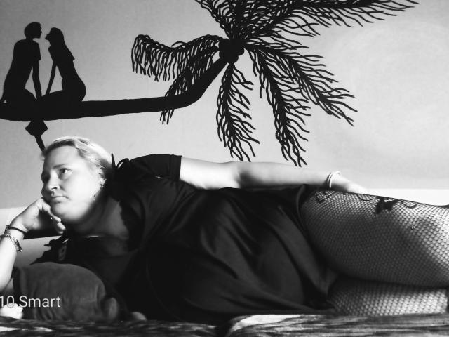 KleinerTeufel82-hot lies on a bed, wearing a black top and fishnet pants, with a palm tree wall decal and silhouetted figures in the background. She's looking off.