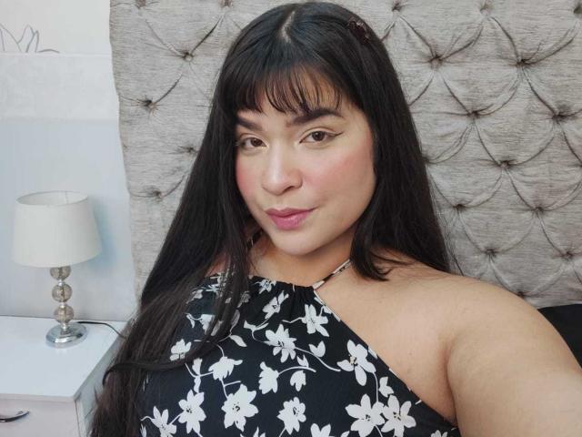 Antonela, mit langen, schwarzen Haaren, Pony und einem schwarzen Top mit Blumenmuster, sitzt neben einer weißen Lampe und zeigt in einem Selfie ihr volles Hinterteil..
