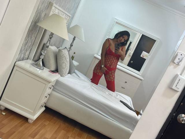 MariaSmith vestindo lingerie vermelha de rede, com uma grande área de nádegas exposta, redonda e nua, com a mão na virilha, em pé em um quarto, tirando uma selfie no espelho..