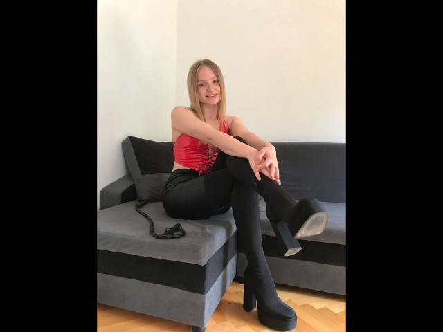 MissNancy-hot with blonde hair, red top, black pants, and boots, sitting on gray sofa, smiling, hands on knees, black lace garment on her right.