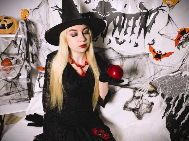 Beatrice, una mujer con cabello rubio largo, que lleva un sombrero de bruja negro, un vestido negro, un collar de cuentas rojas, guantes negros, sosteniendo una manzana roja, con los ojos cerrados, piel pálida, lápiz labial rojo y una mirada sensual..