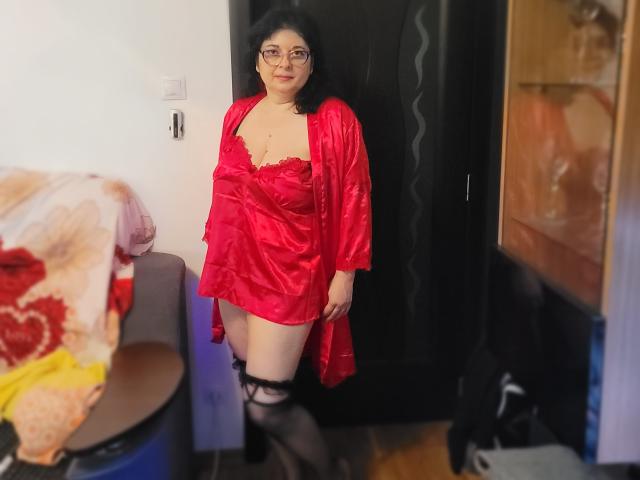 HotAndSexyMature stands in a room wearing a red satin robe, black lace stockings, and glasses, exposing her tits and showing a small pussy. She has curly black hair.