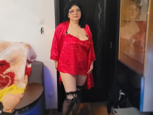 HotAndSexyMature stands in a room wearing a red satin robe, revealing her tits and black lace stockings. She has dark hair, glasses, and a confident expression.