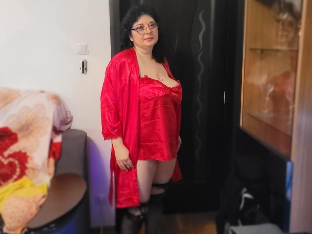 HotAndSexyMature stands in a room wearing a red satin robe, exposing her large tits in a red lingerie top, black thigh-highs, and a slightly open robe. She's looking down.