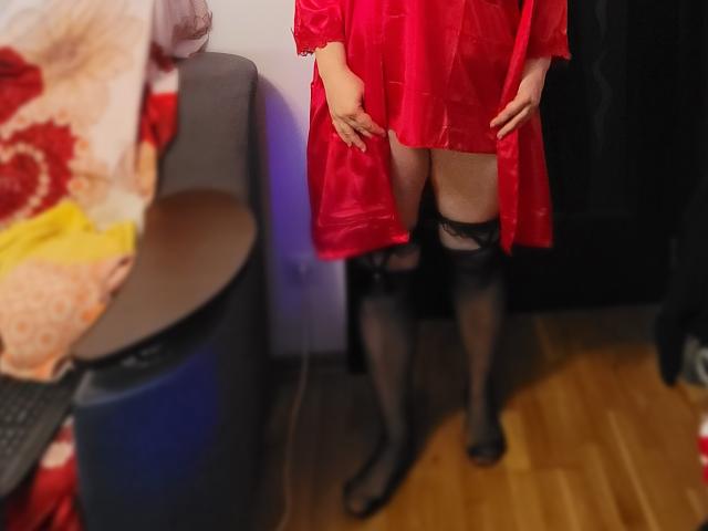 HotAndSexyMature in a red dress, black lace thigh-highs, and black heels, standing with hands on thighs, showing her ass. Hardwood floor, chair, and colorful blanket in background.