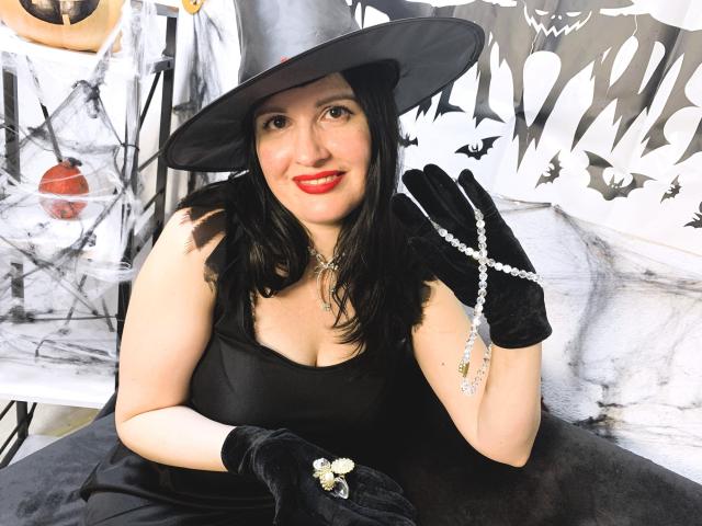 TayaBoret with black dress, black hat, black gloves, red lipstick, holding beaded necklace, showing off tits, sitting, smiling, pale skin, black wavy hair, witchy vibe.