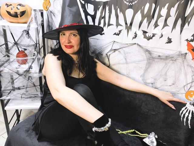 TayaBoret in a black witch hat, red ribbon, black dress, and black boots, sitting with legs up, showing pale skin, red lipstick, black hair, and a bracelet.