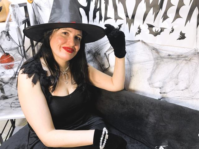 TayaBoret with red lips, black dress, and witch hat, holding hat, wearing black gloves, sitting on gray couch, smiling. Boobs, black hair, pale skin, cute.