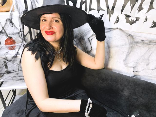 TayaBoret, with red lips, black dress, and hat, sits on a black couch, wearing black gloves, holding her hat, showing her pale skin and black hair, in a Halloween vibe.